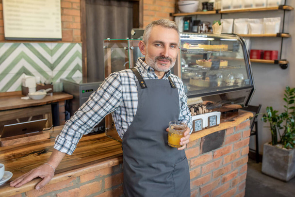 Mid aged man in apron standing near cafe counter with a glass of juice in hand - Foto, immagini