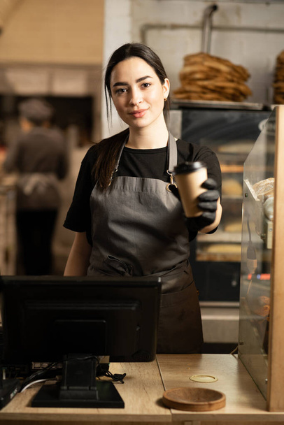 happy saleswoman with paper coffee cup giving americano coffee in vegan cafe. small business, takeaway food, people and service concept - Фото, зображення