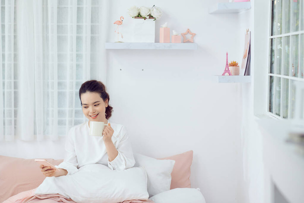 Молода жінка в білому розслабляє і п'є чашку гарячої кави або чаю, використовуючи смартфон на ліжку в спальні
. - Фото, зображення