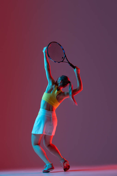 Full length portrait of young woman playing tennis isolated on dark background in neon. Healthy lifestyle. The practicing, fitness, sport, exercise concept. The female model in motion or movement - Fotografie, Obrázek