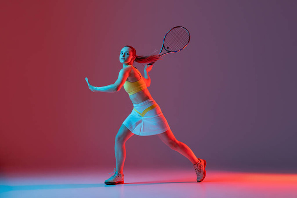 Full length portrait of young woman playing tennis isolated on dark background in neon. Healthy lifestyle. The practicing, fitness, sport, exercise concept. The female model in motion or movement - 写真・画像