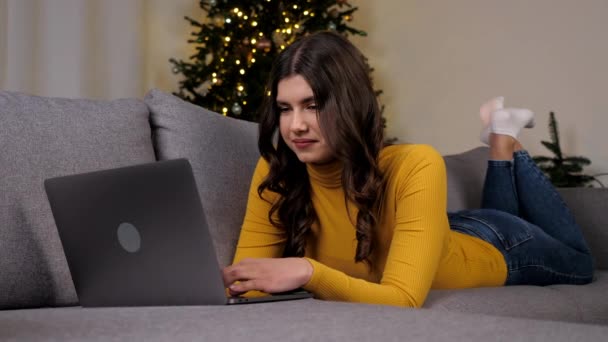 Mosolygó fiatal nő használja laptop online csevegés a legjobb barátok a háttér karácsonyfa. Pozitív gyönyörű női távolság működik számítógép gépelés szöveg billentyűzeten fekvő kanapén otthon - Felvétel, videó