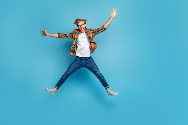 Photo of funky handsome good mood guy open mouth dressed plaid shirt jumping impressed hands to side isolated on blue color background. - Фото, изображение