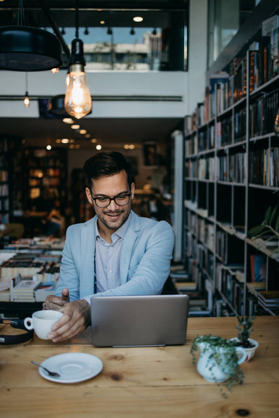 Jeune homme beau assis dans une librairie moderne, lire et utiliser son ordinateur portable ou ordinateur portable. - Photo, image