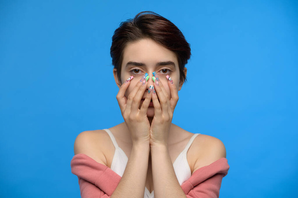 Skinny cute teenage girl hiding her face with both hands playing peek a boo, at blue background - Foto, imagen