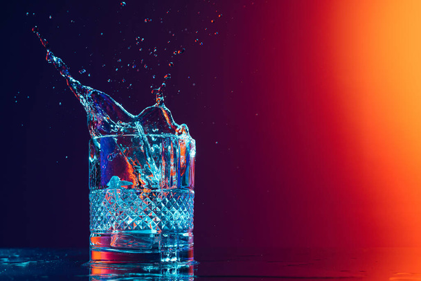 Closeup glass with ice cubes and water splashes standing on mirror surface over gradient black orange background in neon light. Concept of art, beauty, drinks. Copy space for ad - Фото, изображение