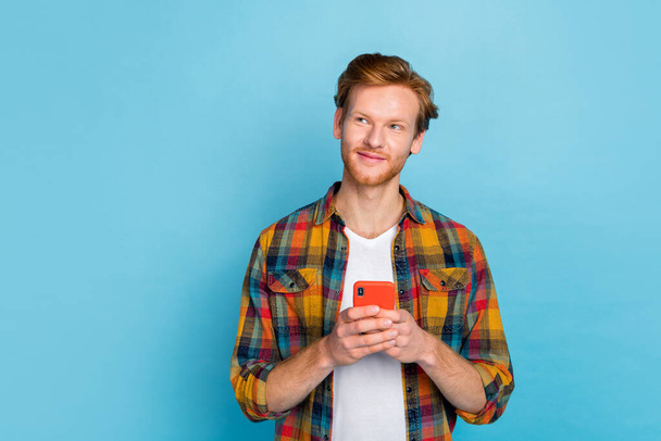 Photo of handsome guy with red hair wear checkered shirt look at empty space holding smartphone isolated on blue color background. - Foto, Imagem