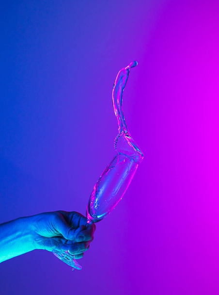 Greeting toast. Authentic hand pouring pure liquid from wine or champagne glass over bright blue-pink gradient background in neon light. Art, drinks, creativity - Foto, Imagen