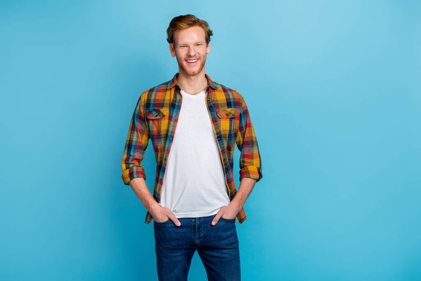 Photo of cheerful positive handsome guy with ginger hairstyle wear checkered shirt arms in pockets isolated on blue color background. - Foto, imagen