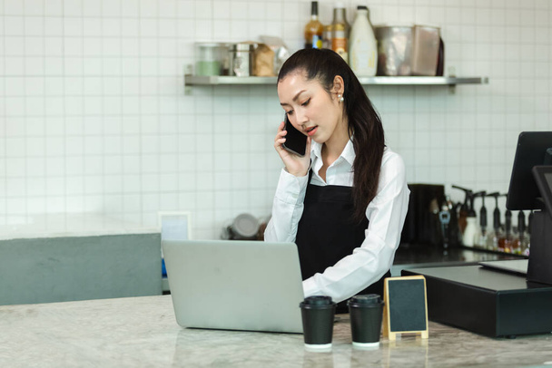 asian young women barista wear apron talking telephone and using laptop computer at coffee shop. female staff standing at counter receive customer orders in cafe. business coffee store concept. - Photo, Image