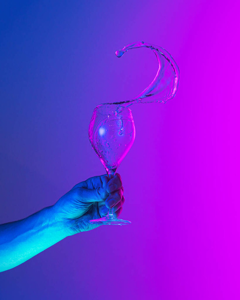Flying fluid. Authentic hand pouring pure water splash from wine or champagne glass over bright blue-pink gradient background in neon light. Art, drinks, creativity - 写真・画像