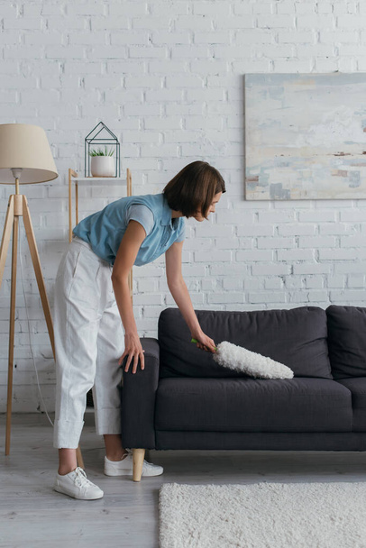 side view of brunette woman cleaning couch in living room at home - Photo, Image