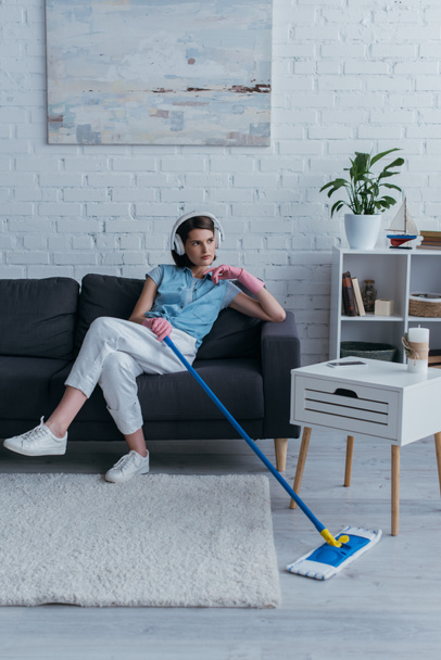 woman holding mop while sitting on couch and listening music in wireless headphones - Photo, Image