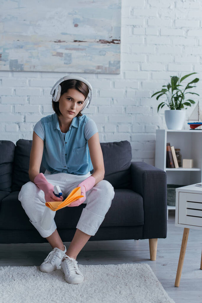 young woman in wireless headphones and rubber gloves sitting on couch and spraying cleanser on rag - Photo, image