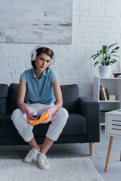 молода жінка в бездротових навушниках і гумових рукавичках сидить на дивані і розпилює засоби для чищення на ганчірці
 - Фото, зображення