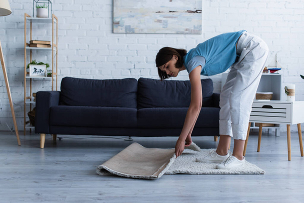 вид збоку молодої жінки, що рухає підлоговий килим у вітальні під час прибирання будинку
 - Фото, зображення