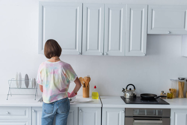 Вид ззаду жінки, що стоїть біля тарілок і раковини на кухні
  - Фото, зображення