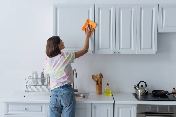 Πλευρική άποψη της νεαρής γυναίκας καθαρισμού λευκό ντουλάπι στην κουζίνα  - Φωτογραφία, εικόνα