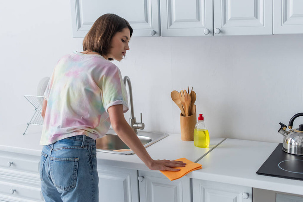 Side view of brunette woman cleaning worktop near sink in kitchen  - Foto, Imagem