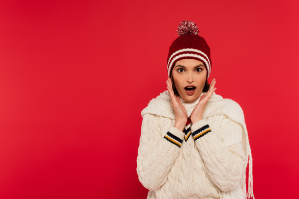 Shocked woman in knitted clothes and hat looking at camera isolated on red  - Photo, image