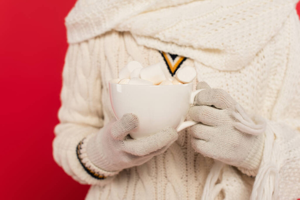 Cropped view of woman in knitted clothes and gloves holding cup with marshmallows isolated on red  - Fotoğraf, Görsel