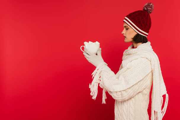 Side view of woman in warm clothes holding cup with marshmallows isolated on red  - 写真・画像