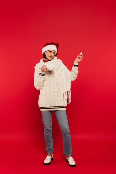 Full length of stylish woman in santa hat holding cup and marshmallow on red background - Foto, immagini