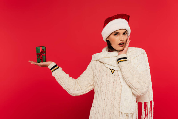 Shocked woman in santa hat holding gift box isolated on red  - Valokuva, kuva