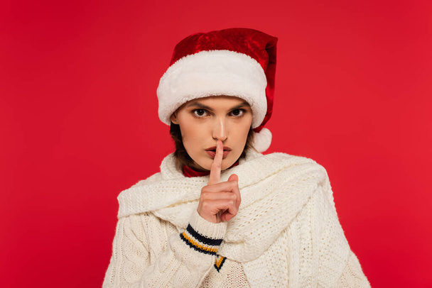 Young woman in santa hat and knitted scarf showing secret gesture isolated on red  - Fotoğraf, Görsel