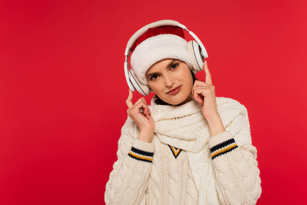 Νεαρή γυναίκα με καπέλο santa και ζεστά ρούχα χρησιμοποιώντας ακουστικά που απομονώνονται στο κόκκινο  - Φωτογραφία, εικόνα