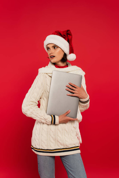 Молода жінка в капелюсі Санта і светрі тримає ноутбук ізольовано на червоному
  - Фото, зображення