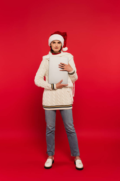 Full length of woman in warm clothes and santa hat holding laptop on red background - Photo, image