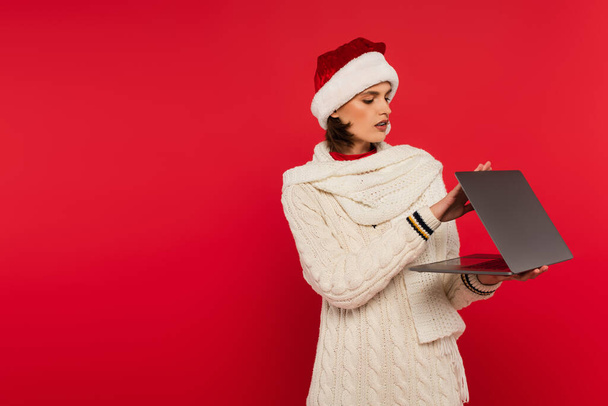 Woman in santa hat and warm scarf holding laptop isolated on red  - Valokuva, kuva
