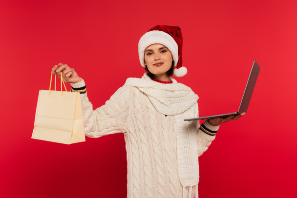 ευχαριστημένη νεαρή γυναίκα με καπέλο santa και πλεκτό πουλόβερ κρατώντας φορητό υπολογιστή και τσάντα για ψώνια σε κόκκινο - Φωτογραφία, εικόνα