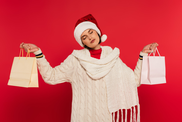 young woman in santa hat and knitted sweater holding shopping bags on red - Foto, Imagem