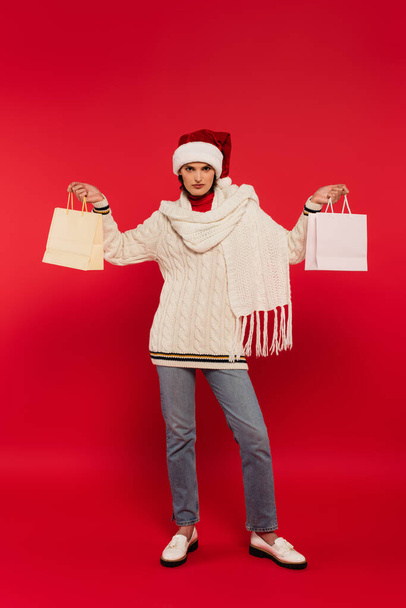 full length of young woman in santa hat and knitted sweater holding shopping bags on red - Фото, зображення