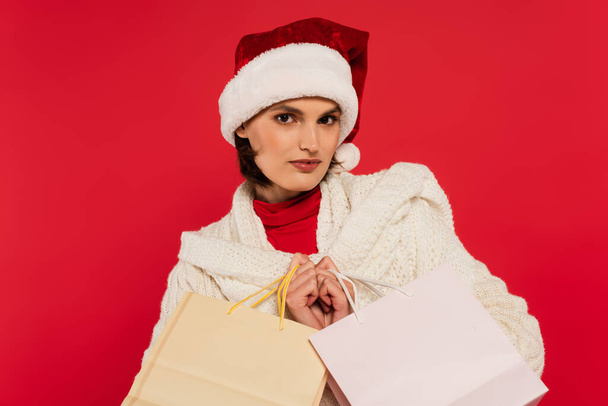 young woman in santa hat and knitted sweater holding shopping bags isolated on red - Fotó, kép