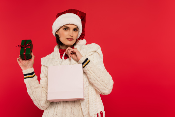 young woman in santa hat holding shopping bag and gift box with bow isolated on red - Fotoğraf, Görsel