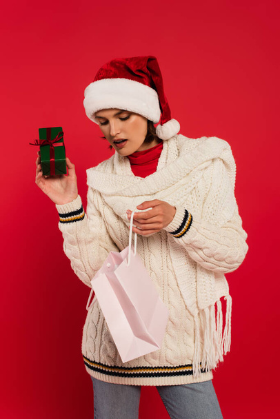 amazed young woman in santa hat holding shopping bag and gift box with bow isolated on red - Fotografie, Obrázek