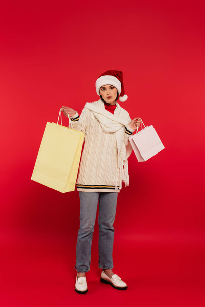 full length of surprised young woman in santa hat and winter outfit holding shopping bags on red - Zdjęcie, obraz