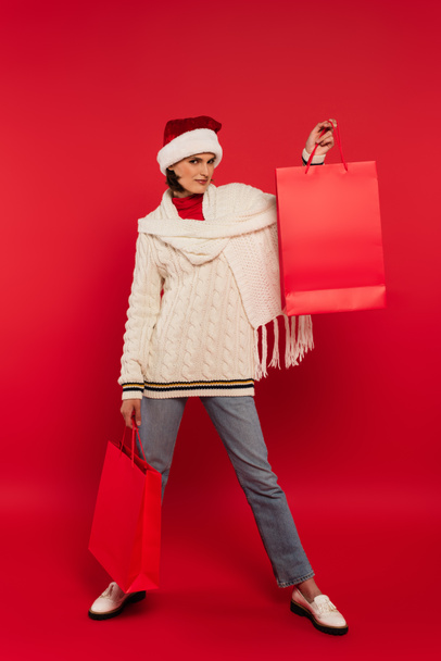 full length of young woman in santa hat and winter outfit holding shopping bags on red - Fotoğraf, Görsel