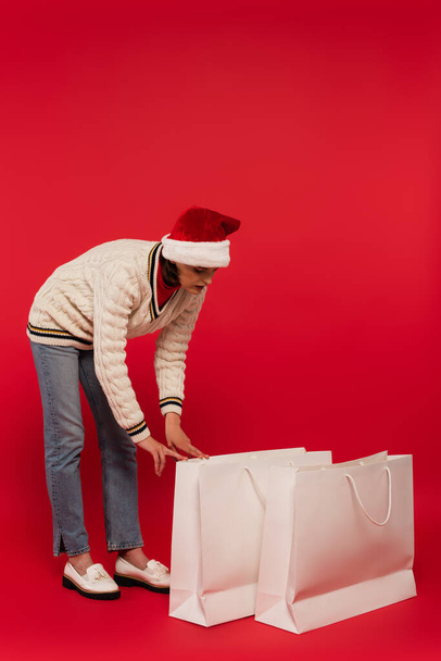 full length of young woman in santa hat looking at shopping bags on red - Фото, изображение