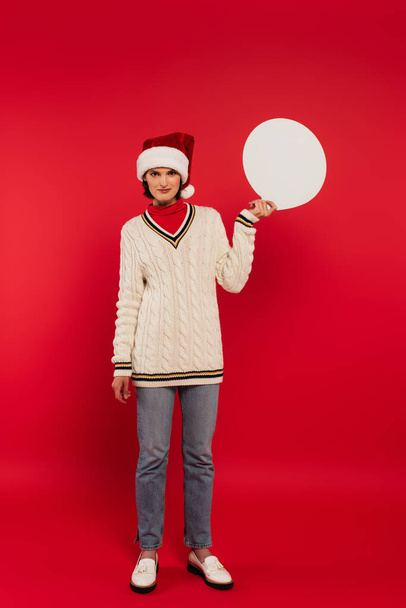 πλήρες μήκος της νεαρής γυναίκας σε santa καπέλο και πουλόβερ κρατώντας φούσκα ομιλία στο κόκκινο - Φωτογραφία, εικόνα