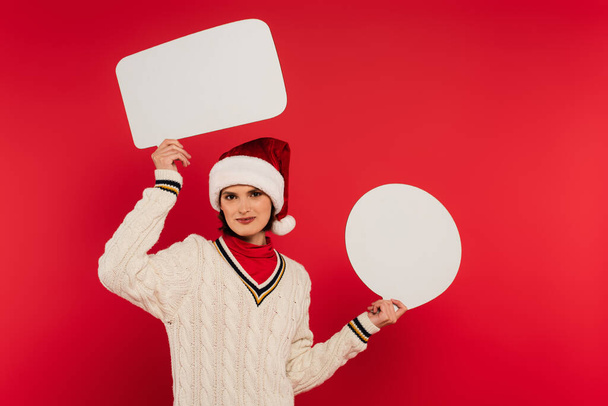 smiling young woman in santa hat and sweater holding speech bubbles isolated on red - Zdjęcie, obraz