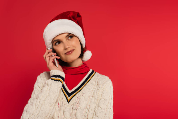 улыбающаяся молодая женщина в шляпе Санты и вязаном свитере разговаривает на смартфоне, изолированном на красном - Фото, изображение