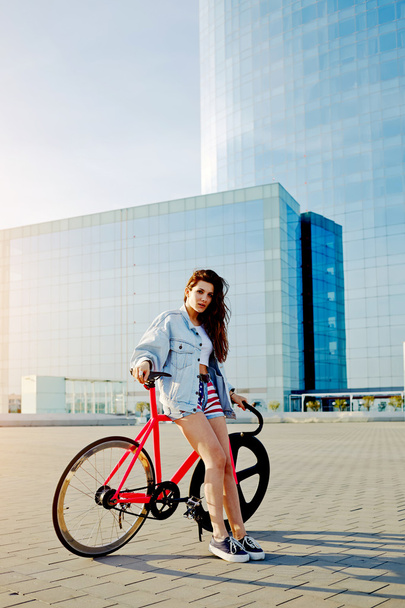 Attractive young woman win fixed gear bike posing outdoors with beautiful skyscraper on background, pretty young brown haired woman standing with her modern pink bicycle in city, stylish hipster girl - Фото, изображение