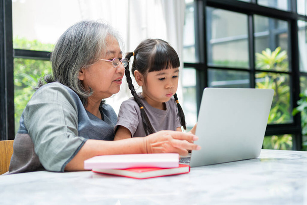 Asyalı büyükanne ve iki torunu evde bilgisayar defteriyle oyun oynuyorlar ve eğleniyorlar. Çevrimiçi eğitim kavramı ve ebeveynlerin bakımı. - Fotoğraf, Görsel