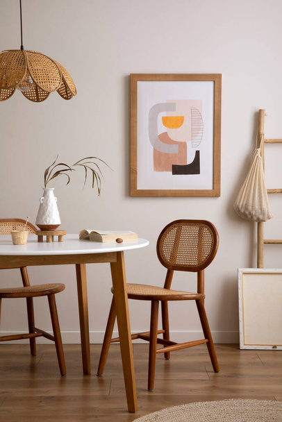 The stylish dining room with round table, rattan chair, wooden commode, pock up poster and kitchen accessories. Beige wall with mock up poster. Home decor. Template.  - Foto, Imagen