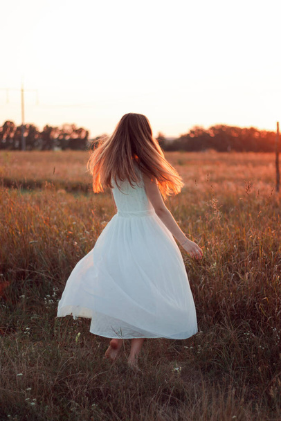 Χαριτωμένο κορίτσι κάνει κύκλους με ένα λευκό φόρεμα στο γήπεδο. Ακτίνες ηλίου. Πορτοκαλί ηλιοβασίλεμα. Έννοια. Μια γυναίκα χορεύει χαρούμενα - Φωτογραφία, εικόνα