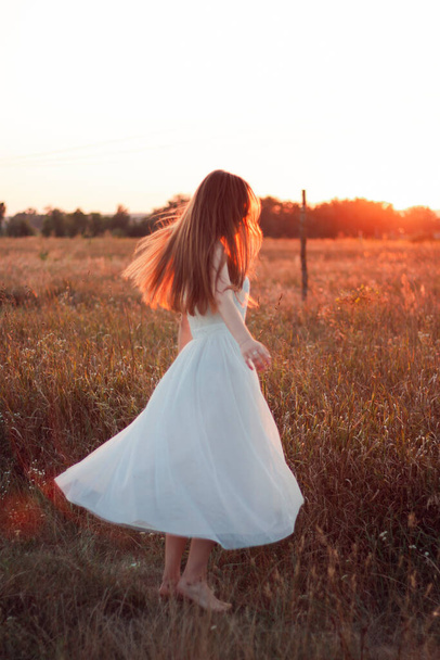 Pretty cute girl is circling in a white dress on the field. Sun rays. Orange sunset. Concept. A woman dances joyfully - Photo, image